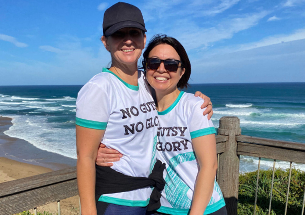 Two women in matching No Gutsy No Glory shirts embrace on a sunny coastal path